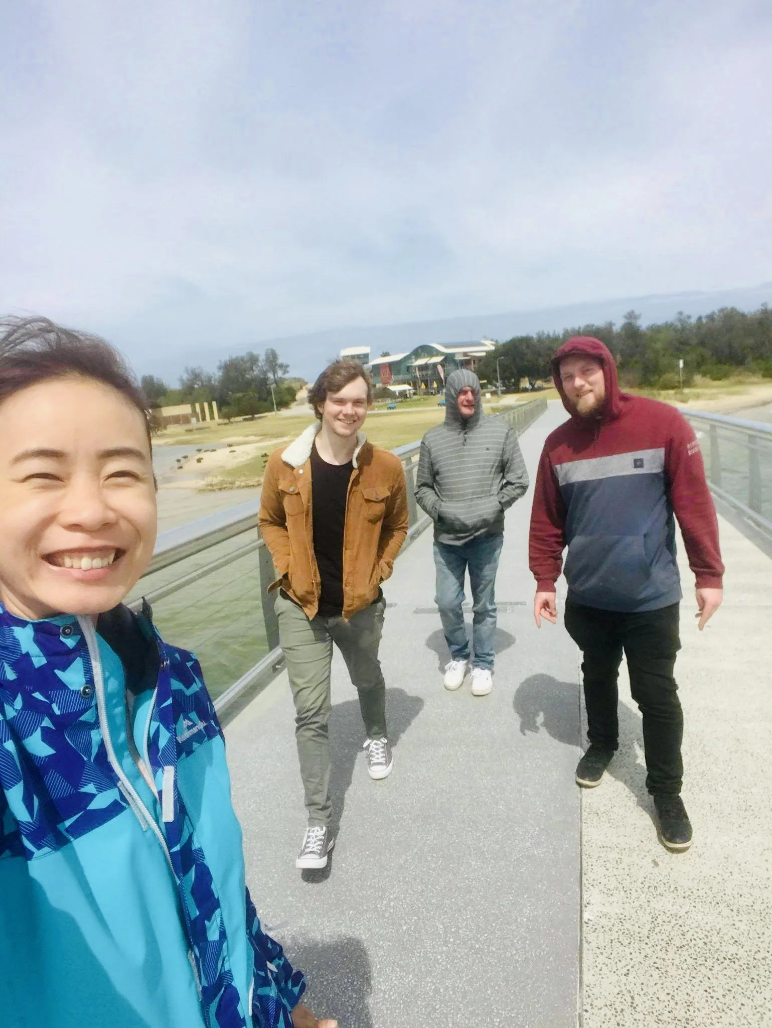 Group of four friends walking on a bridge outdoors, smiling, with a building and trees in the background.