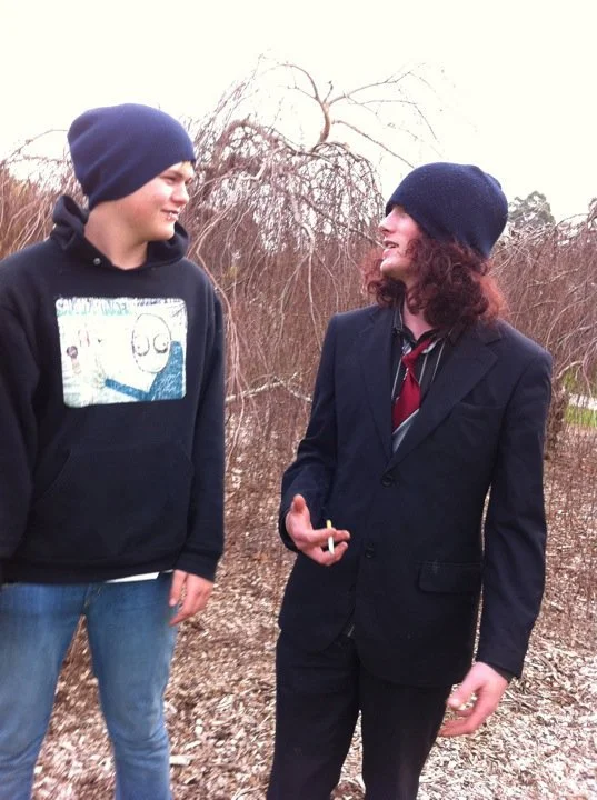Two young men with curly hair and beanies standing outdoors, facing each other and smiling, with leafless bushes in the background.