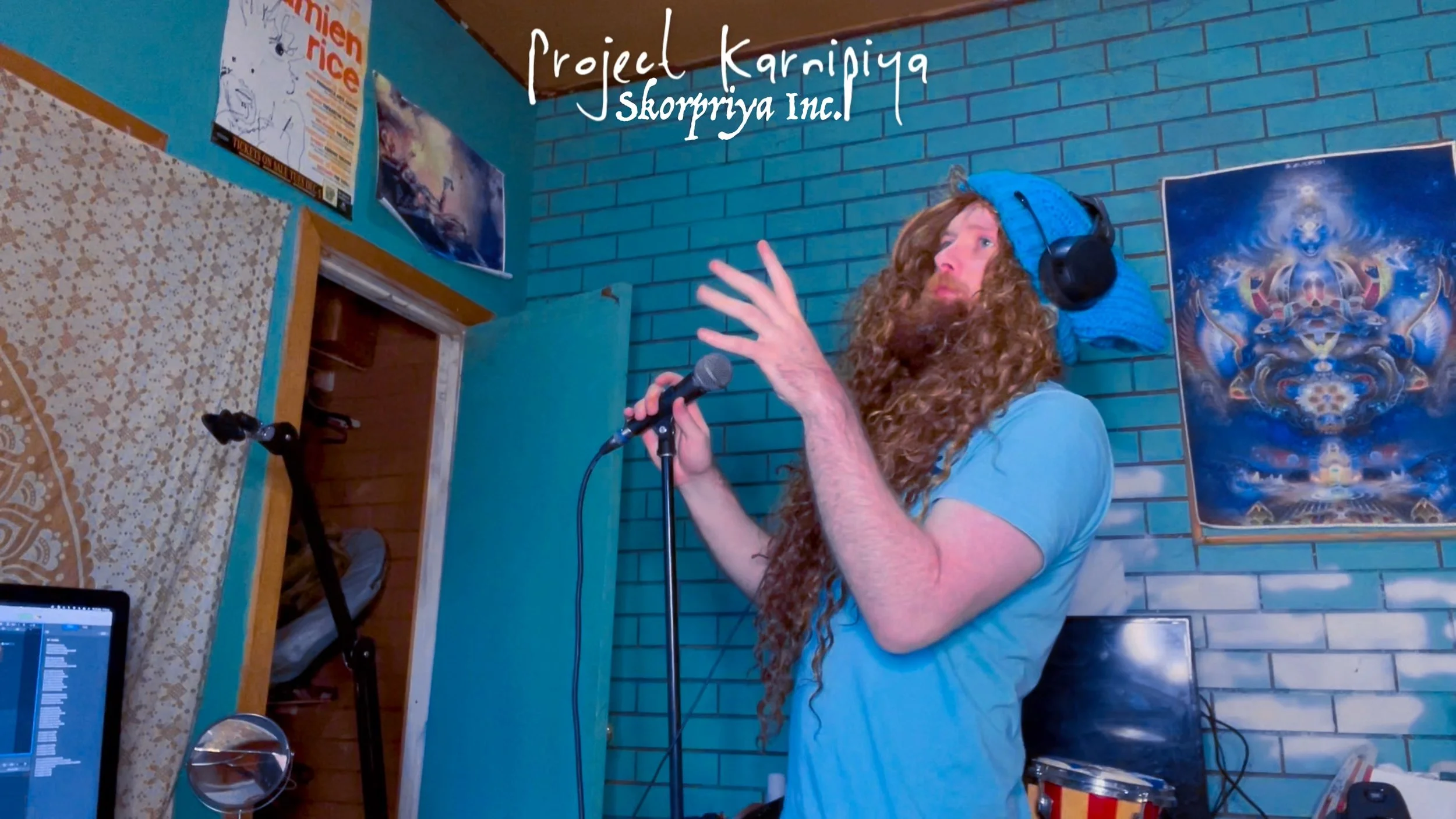 A man with long, curly red hair and a beard wearing headphones and a blue t-shirt, holding a microphone and gesturing with his left hand in a room with blue brick walls and posters, including one with abstract art.