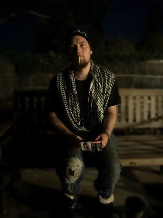 A young man sitting outdoors on a bench at night, wearing a black t-shirt, a checkered scarf, and ripped jeans, holding a lighter and a pack of cigarettes.