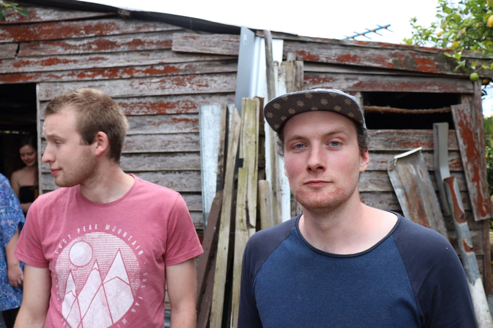Two young men standing outdoors in front of a weathered wooden shed, with one wearing a pink shirt and a person smiling in the background.