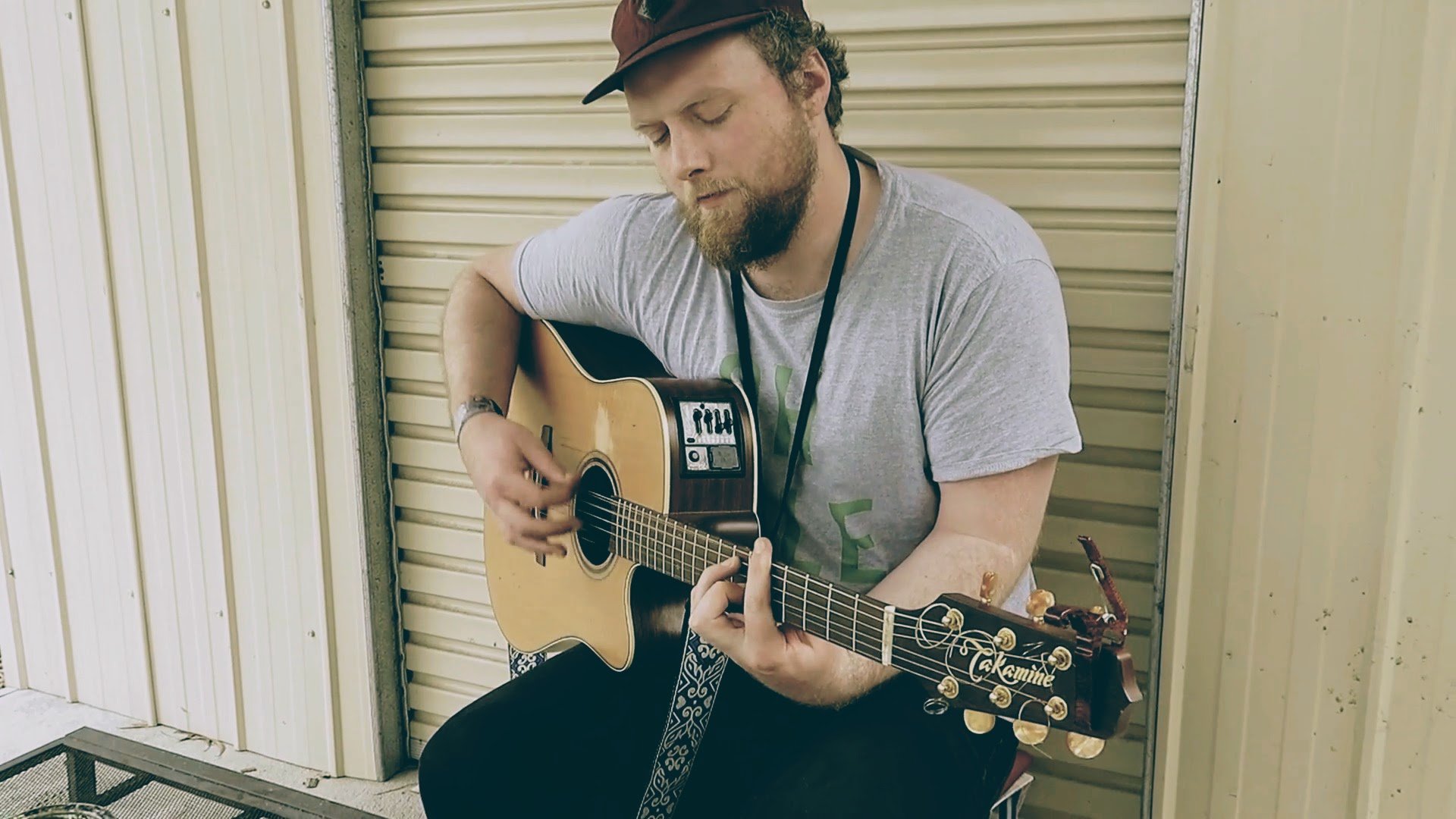 A man with a beard and cap sitting against a beige metal wall, playing an acoustic guitar.