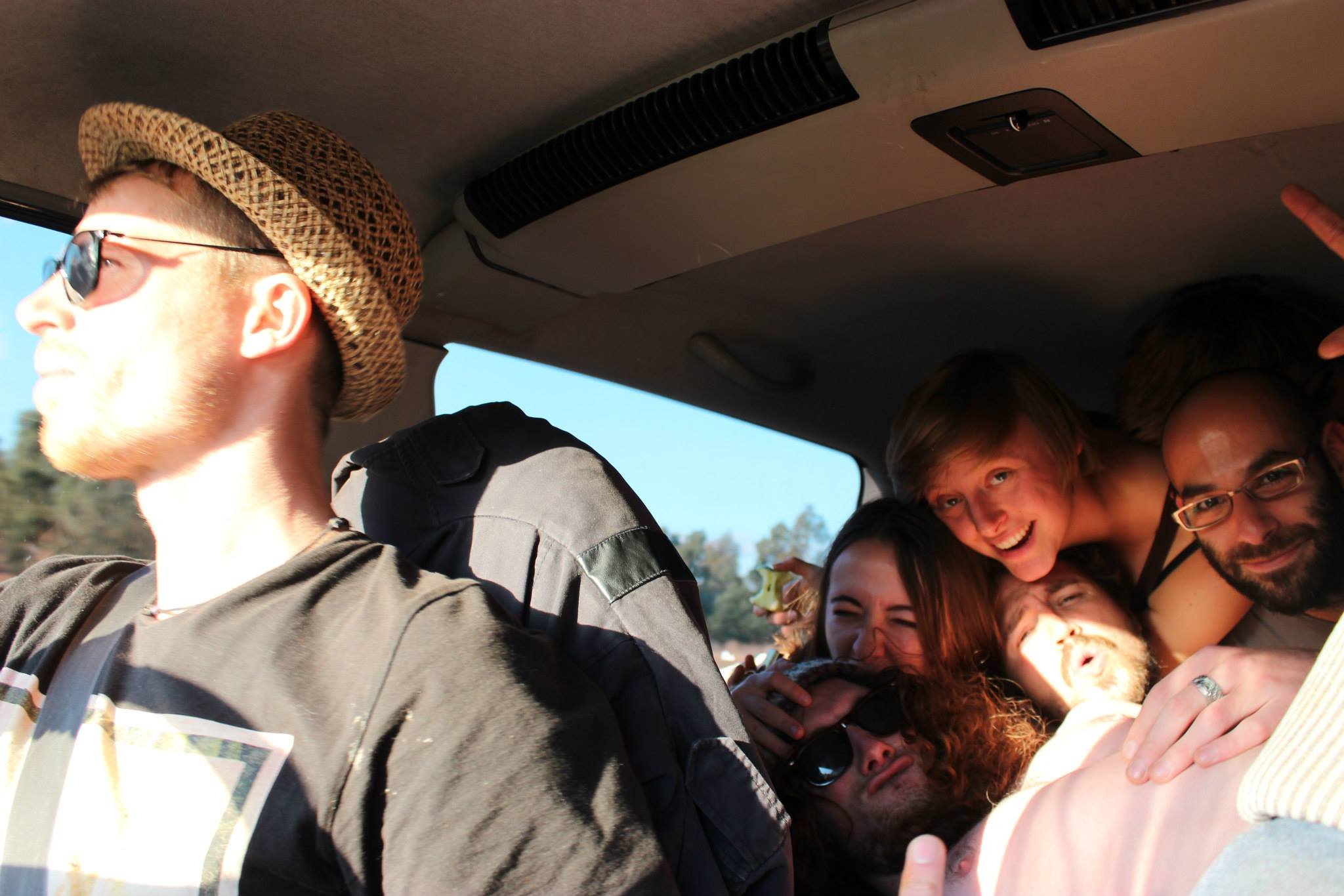 Group of friends sitting close together in a vehicle, some are smiling and posing for the camera, with a man in front driving, wearing sunglasses and a straw hat.