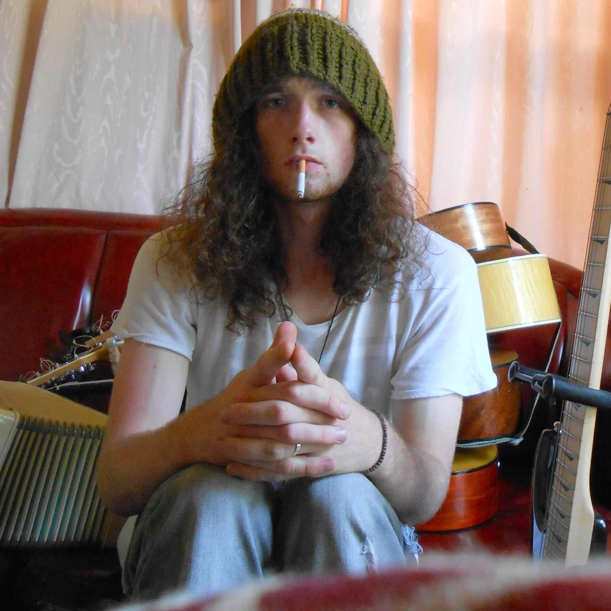 A young man with long curly hair wearing a green knit beanie, white t-shirt, and gray pants, sitting with hands clasped, smoking a cigarette in a room with guitars and curtains in the background.