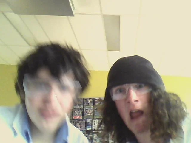 Two young men with curly hair and glasses taking a selfie indoors, with a yellow wall and display shelves in the background.