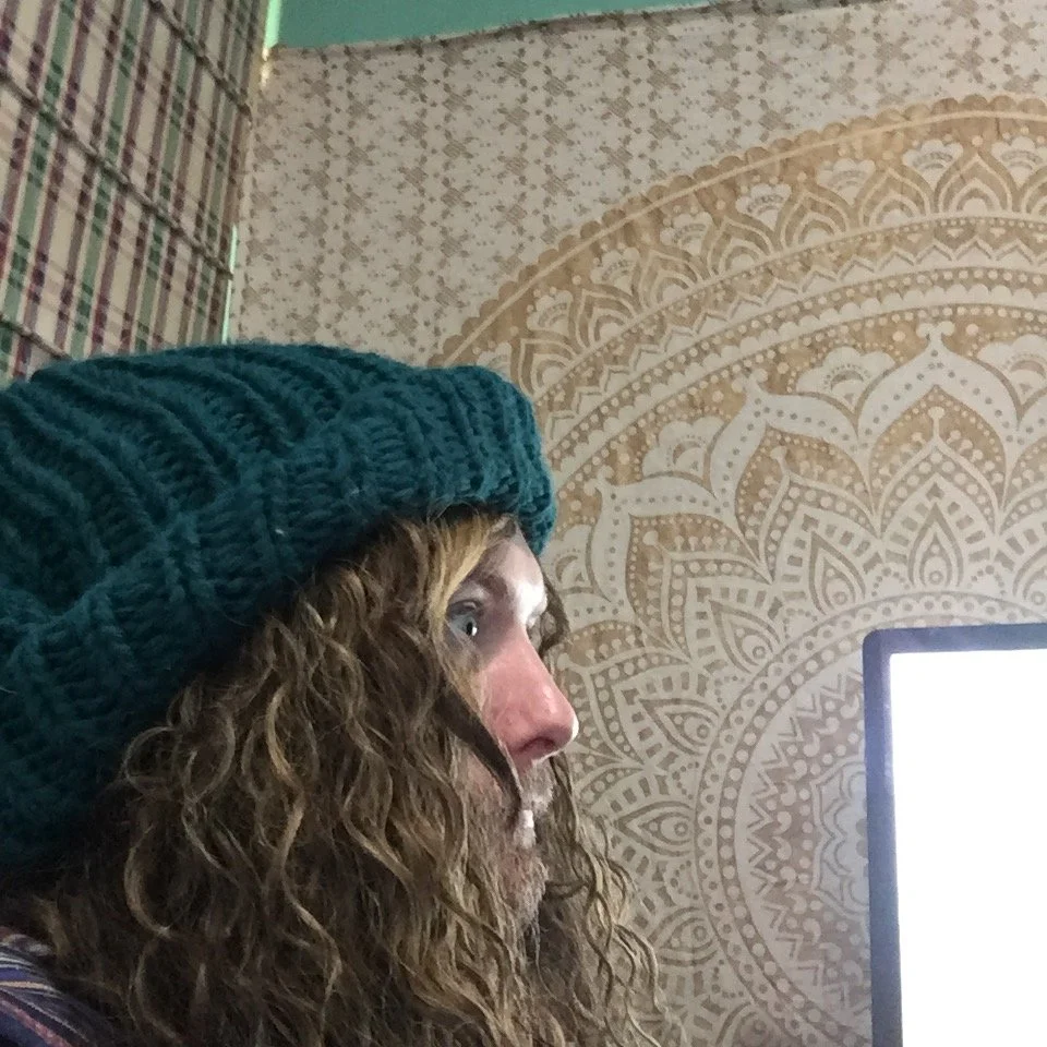 A woman with curly hair wearing a teal knit hat, looking at a computer monitor, in front of a patterned wall with beige and white designs.