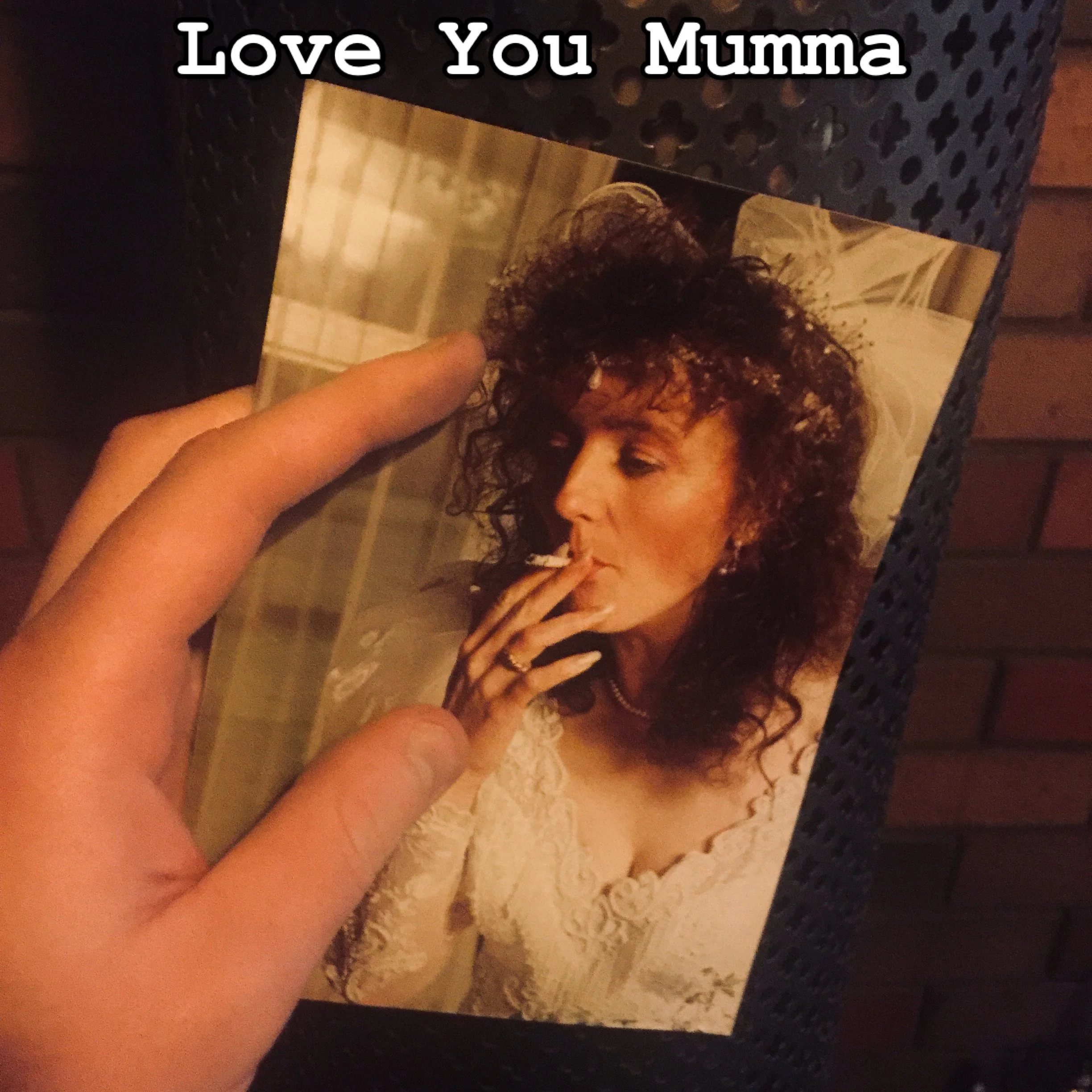 A person holding a photo of a woman with curly hair, smoking a cigarette, dressed in white, with the text 'Love You Mumma' written at the top.