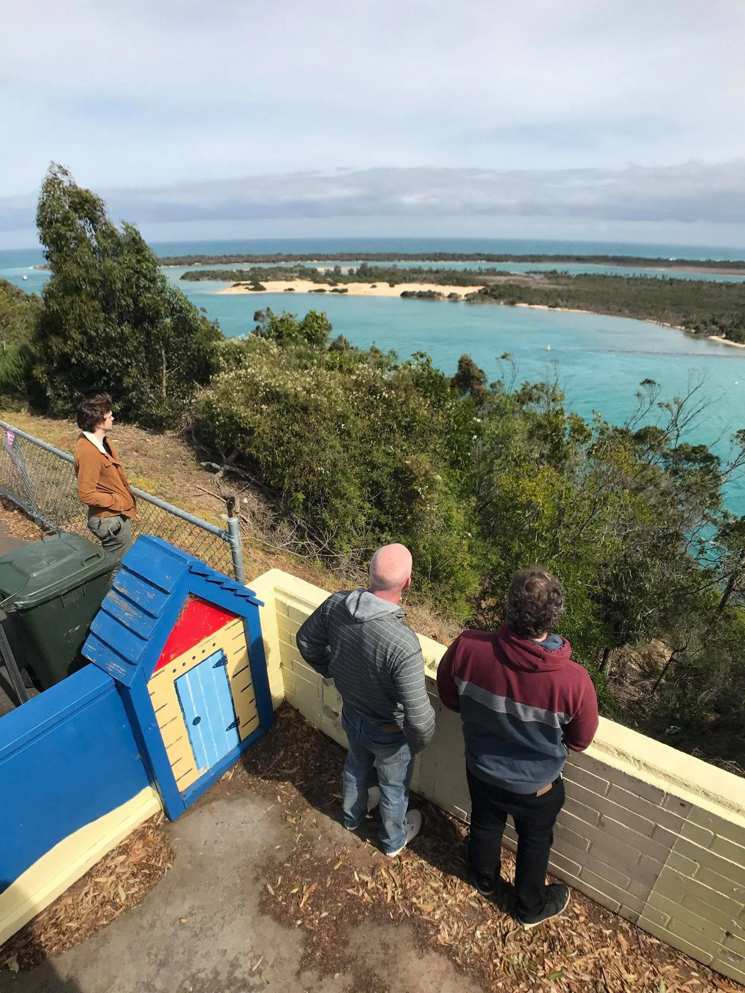 Three people stand on a viewing platform overlooking a coastal landscape with trees, blue water, sandy beaches, and a distant landmass under a partly cloudy sky.