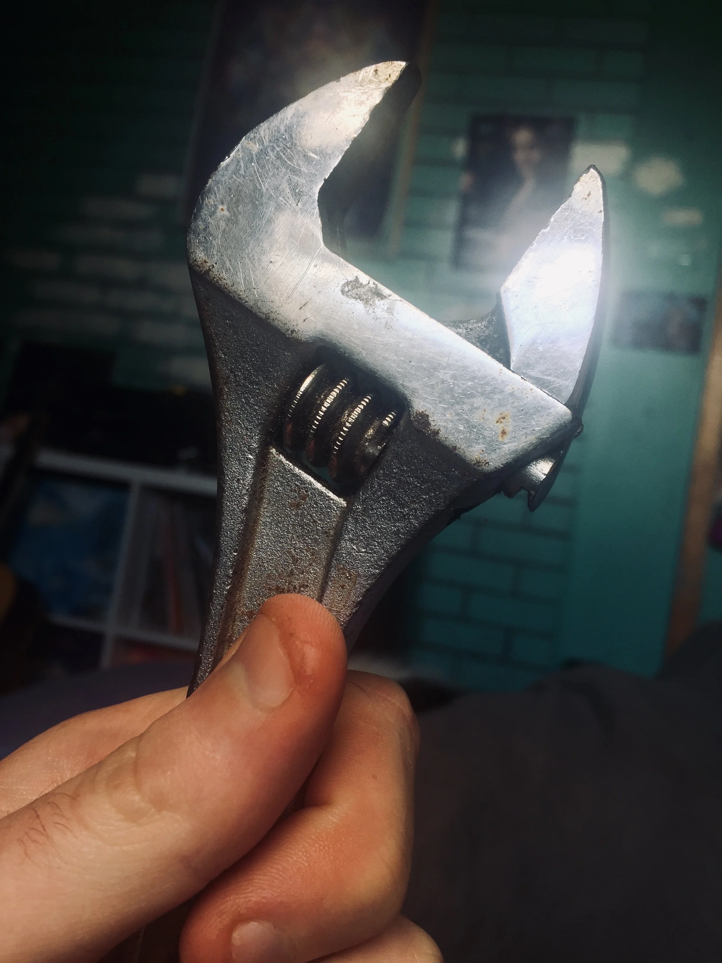 A person holding an adjustable wrench with a light shining across the silver metal surface, against a background of a teal wall with posters or pictures.