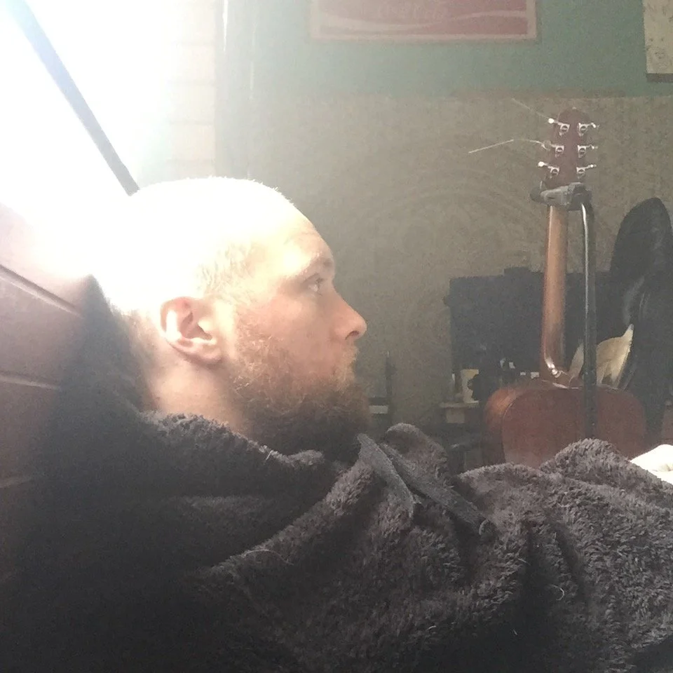 A man with a blond beard and shaved head lying on a bed, looking to the right, with a guitar in the background.