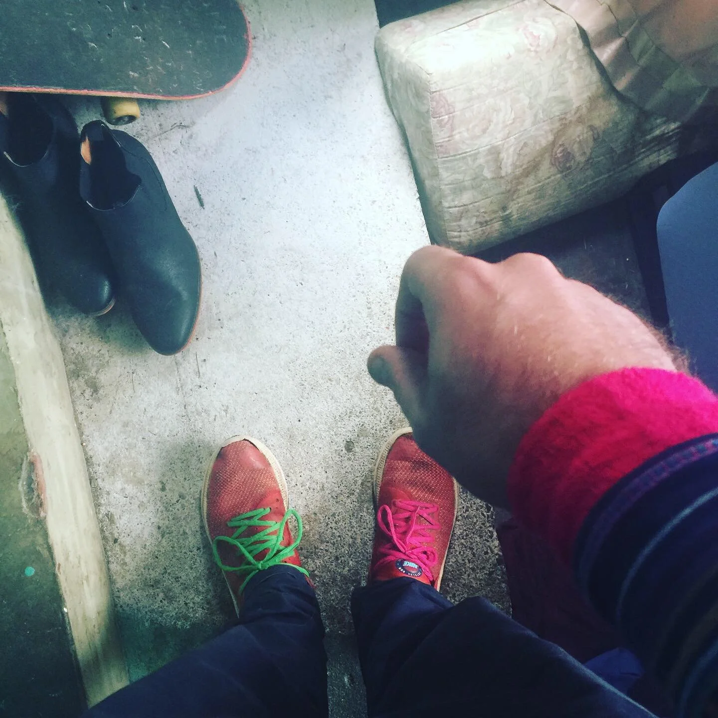 Looking down at a person's colorful sneakers, one with green laces and the other with pink laces, on a concrete floor near a skateboard, black shoes, and a beige armchair.