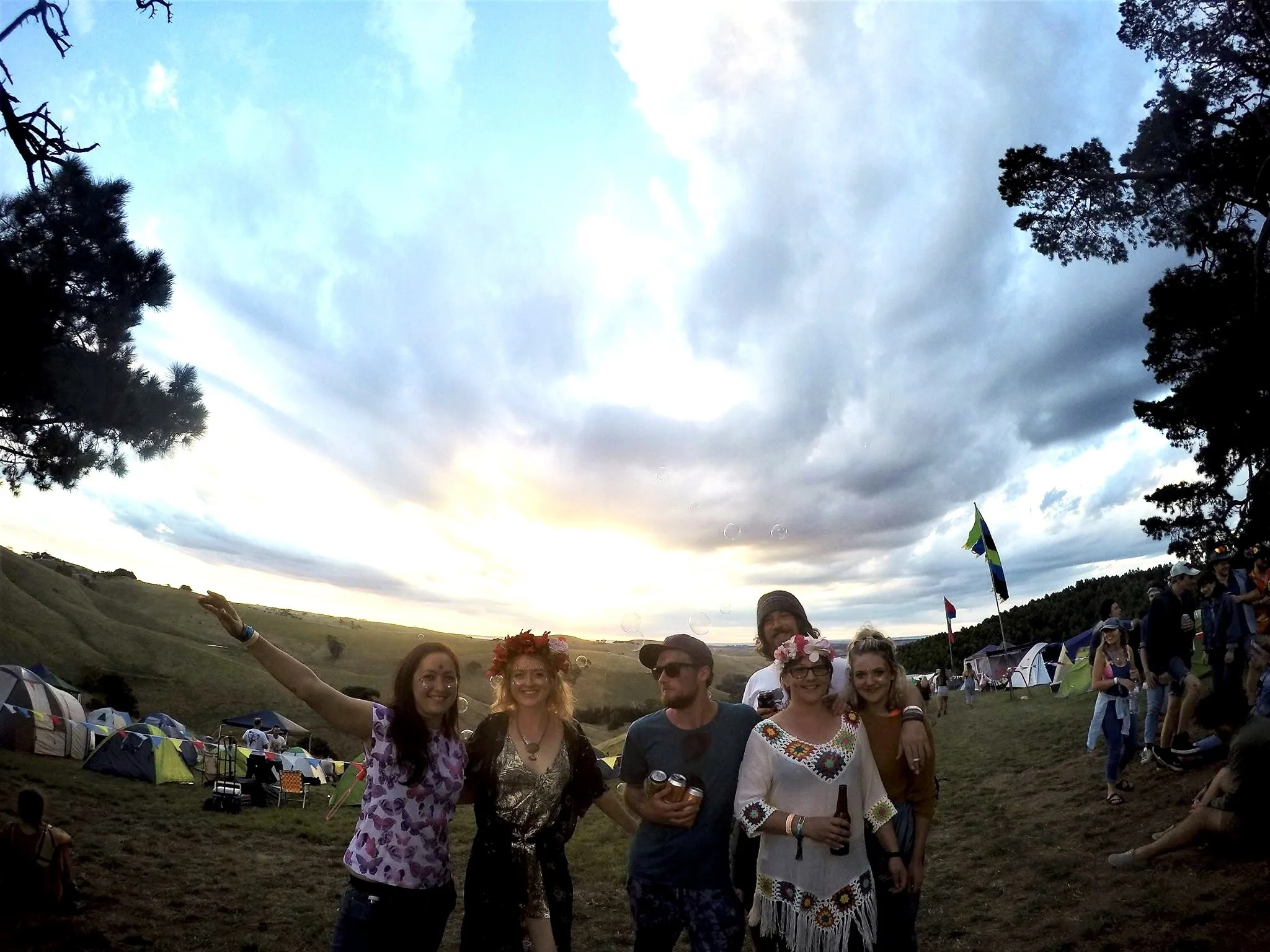 A group of friends at a festival, standing on a grassy hill with tents in the background, during sunset with a partly cloudy sky. Four women and one man are smiling, some wearing flower crowns, and holding drinks.