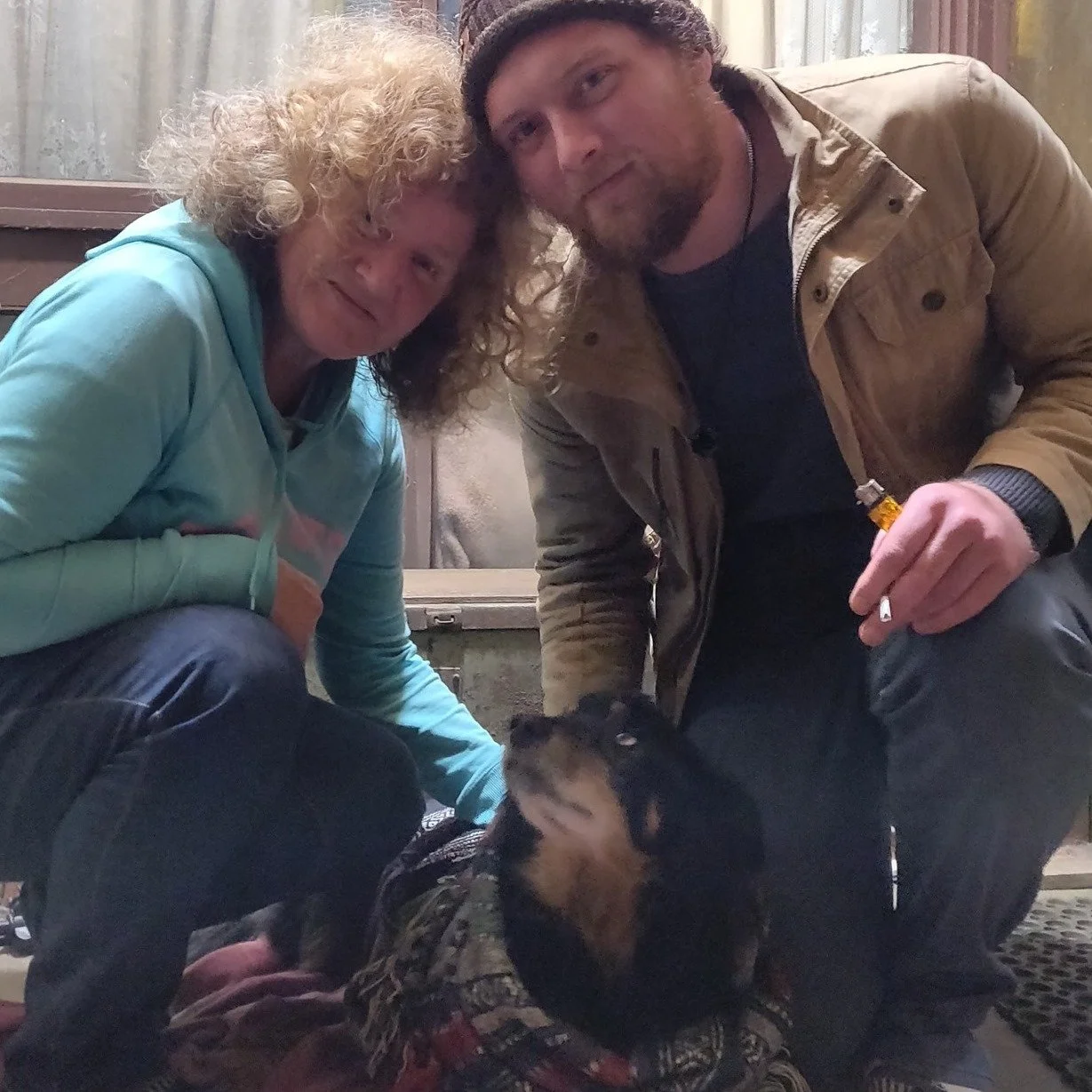 Two people, a woman with curly blonde hair and a man with a beard, are crouching on either side of a black and tan dog. They are outdoors near a wooden building. The woman is wearing a light blue hoodie, and the man is wearing a tan jacket and a bean