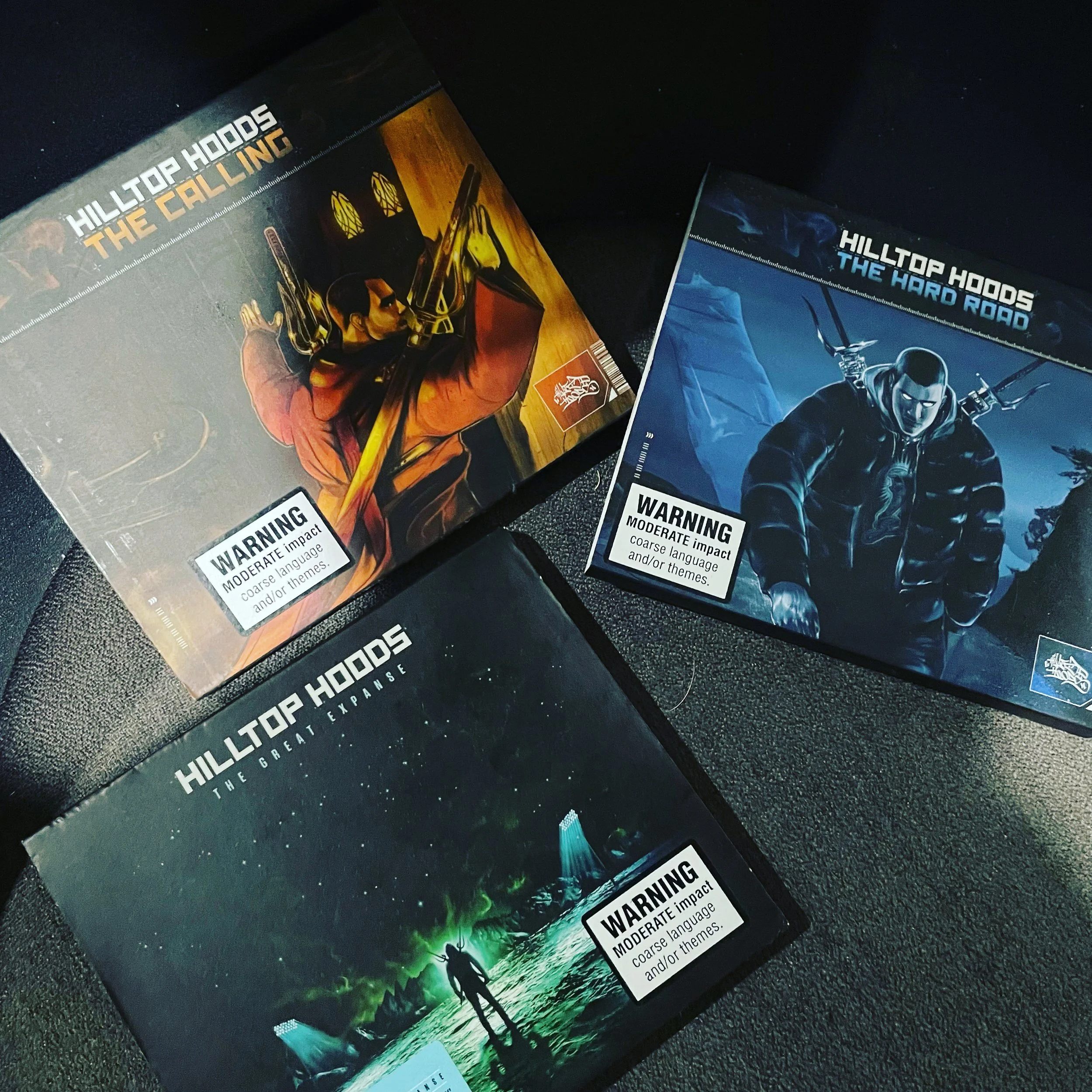 Three tabletop game boxes titled "Hilltop Hoods: The Callling," "Hilltop Hoods: The Hard Road," and "Hilltop Hoods: The Great Expanse," each with cover art depicting animated characters in different scenes.