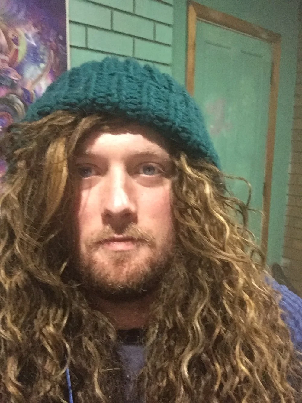 A man with long curly hair and a beard wearing a teal knit beanie, looking at the camera in a room with a green brick wall and posters.