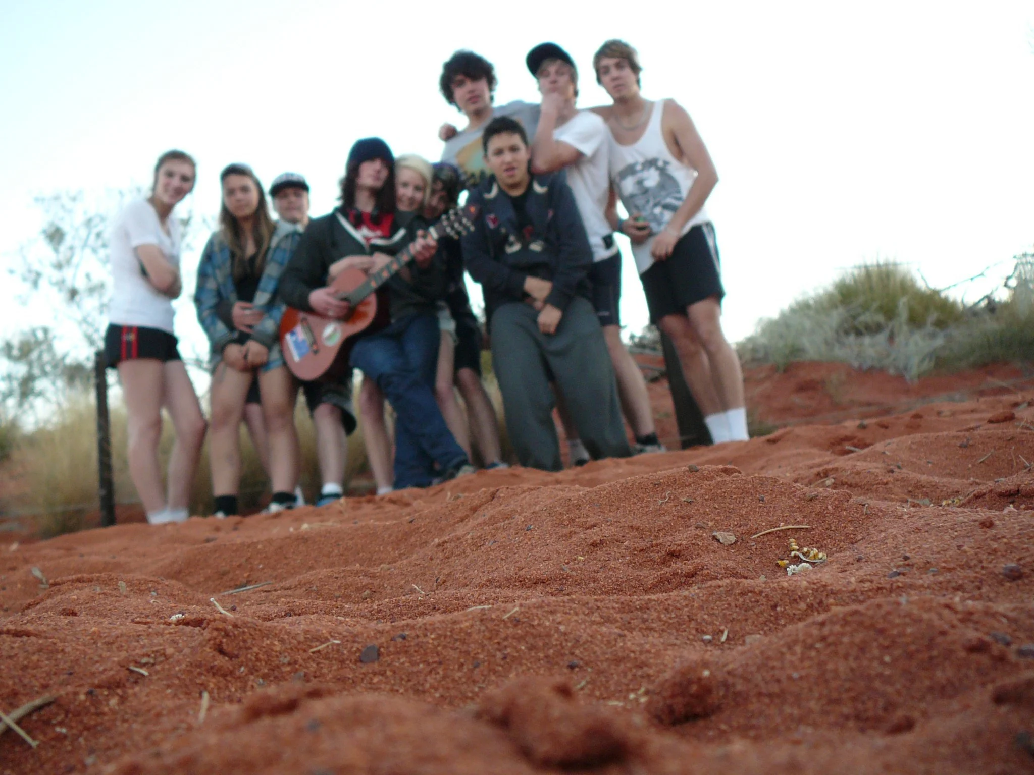 A group of young people standing on red dirt ground outdoors, with some holding musical instruments and others posing for a photo.
