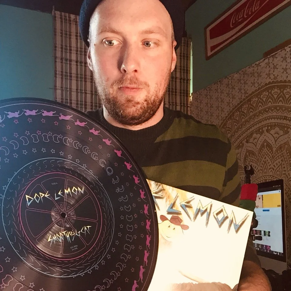 A man with a beard and mustache wearing a dark beanie and a striped shirt holding a vinyl record titled 'Dark Lemon' in a room with a computer monitor, framed artwork, and patterned walls.