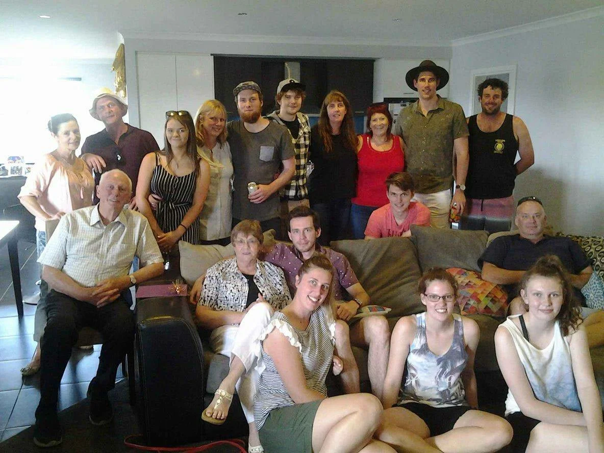 Group of 14 people gathered in a living room for a party or celebration, with some seated on the floor and others standing behind them, smiling at the camera.