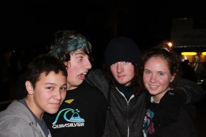 Four teenagers standing close together at night, with three of them smiling and one making a funny face.