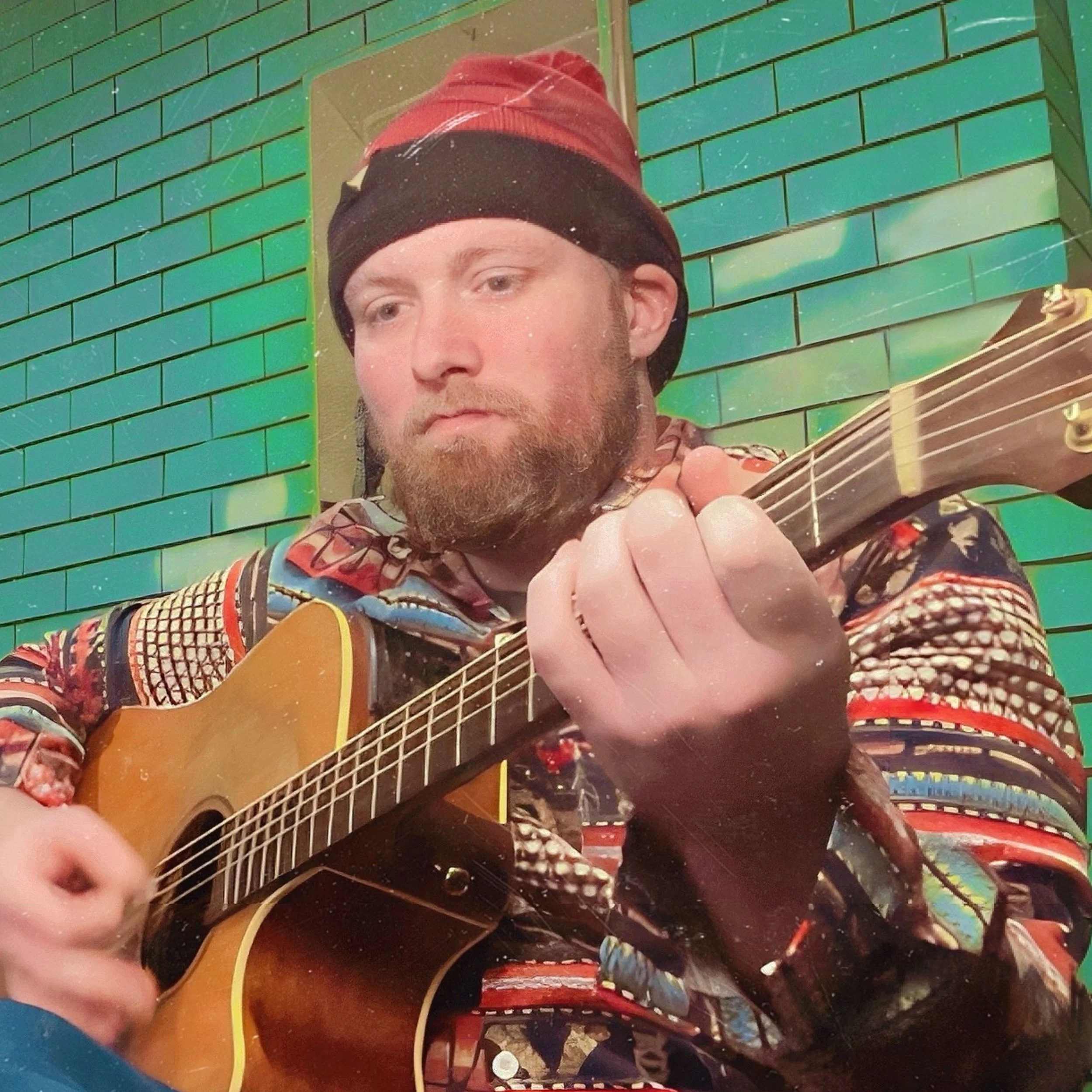 A man with a beard wearing a multicolored jacket and a red and black beanie hat, playing an acoustic guitar in front of a green brick wall.