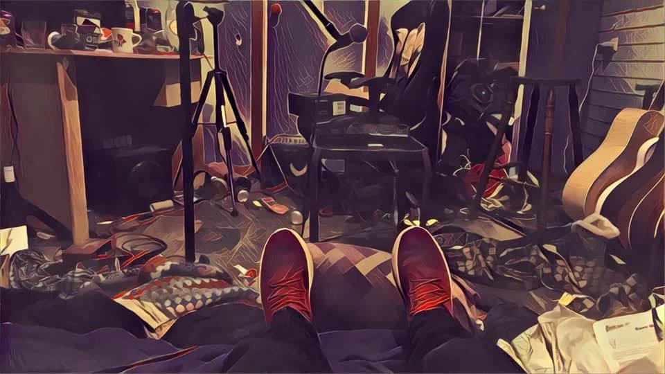 A cluttered room with musical equipment, a guitar, and various electronic devices, viewed from a person's perspective sitting on a bed with red shoes.