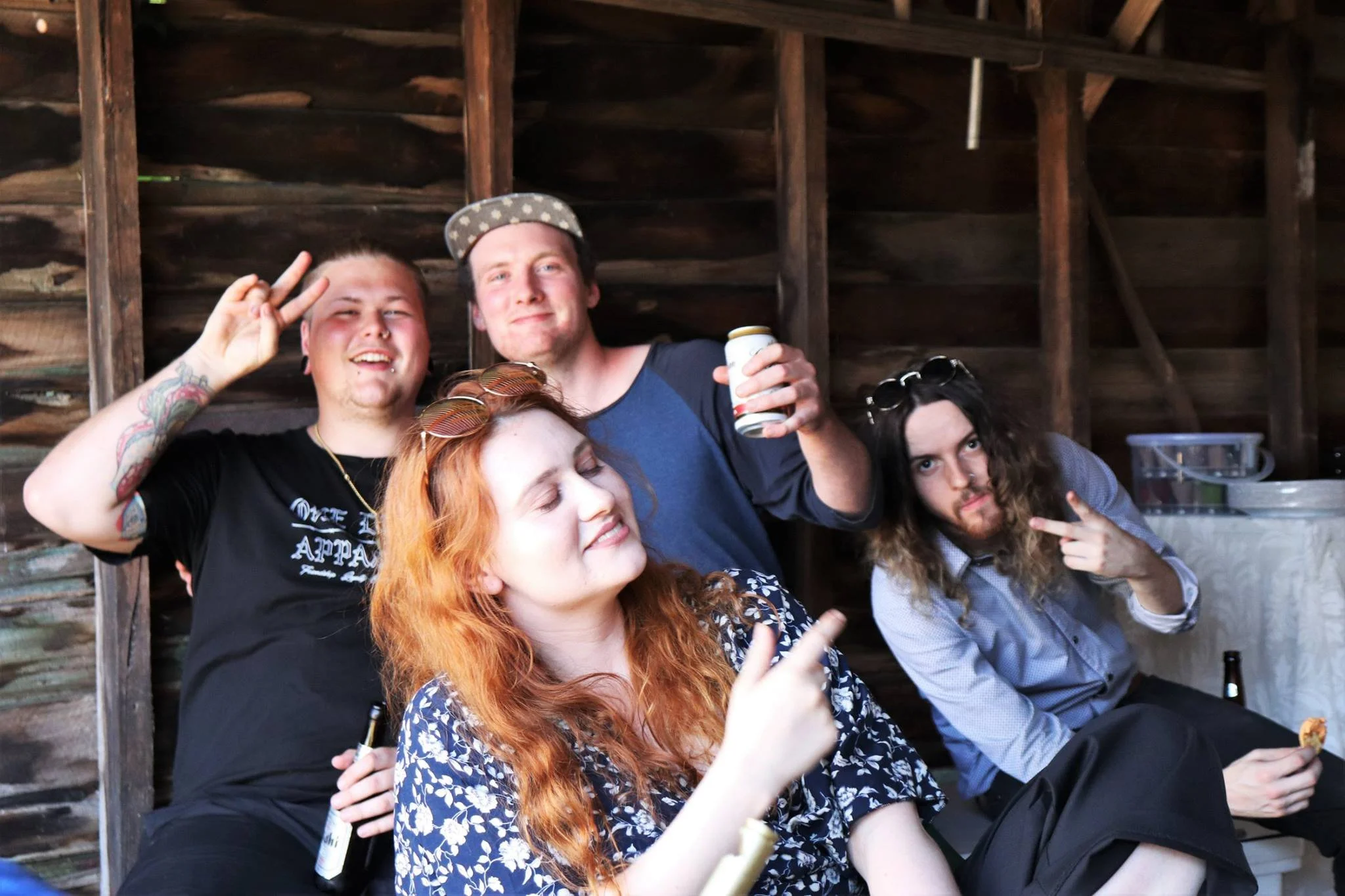 Four friends sitting and standing in a rustic setting, smiling and making peace signs or pointing, with one holding a beer can and another holding a beer bottle.