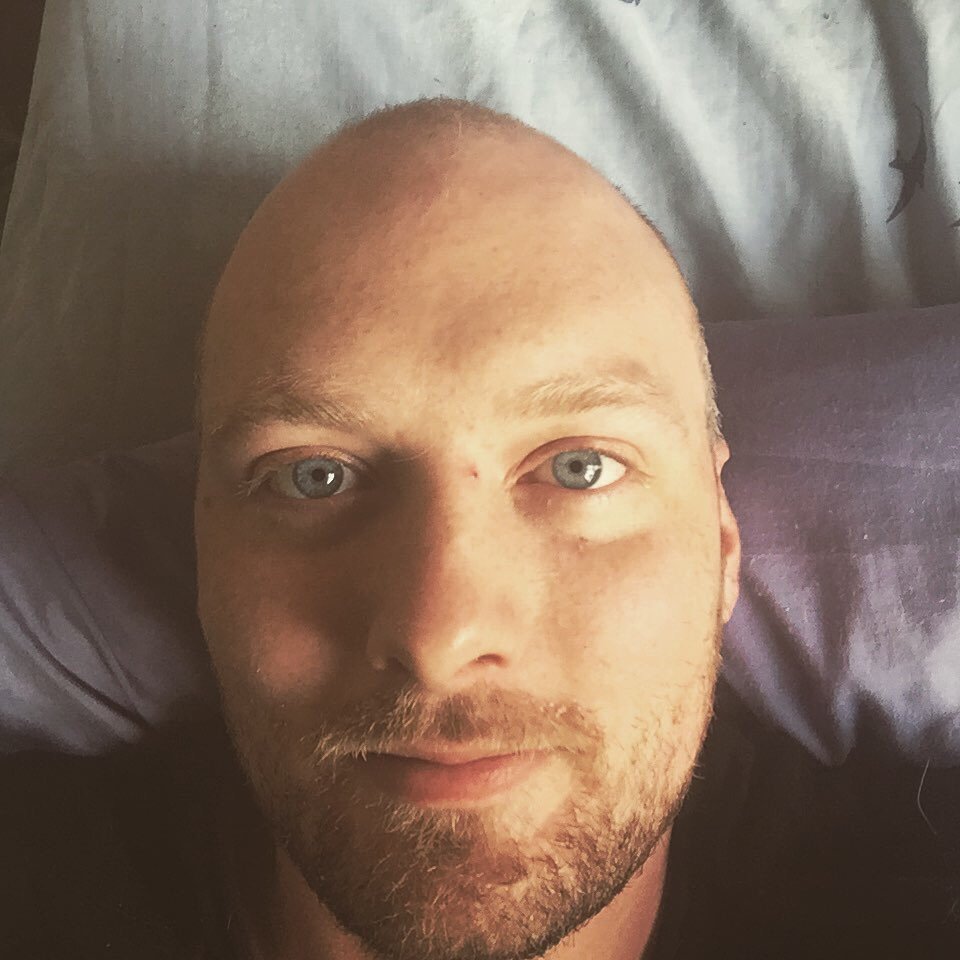 Close-up selfie of a man with a bald head, light skin, blue eyes, and facial stubble, lying on a pillow in bed.
