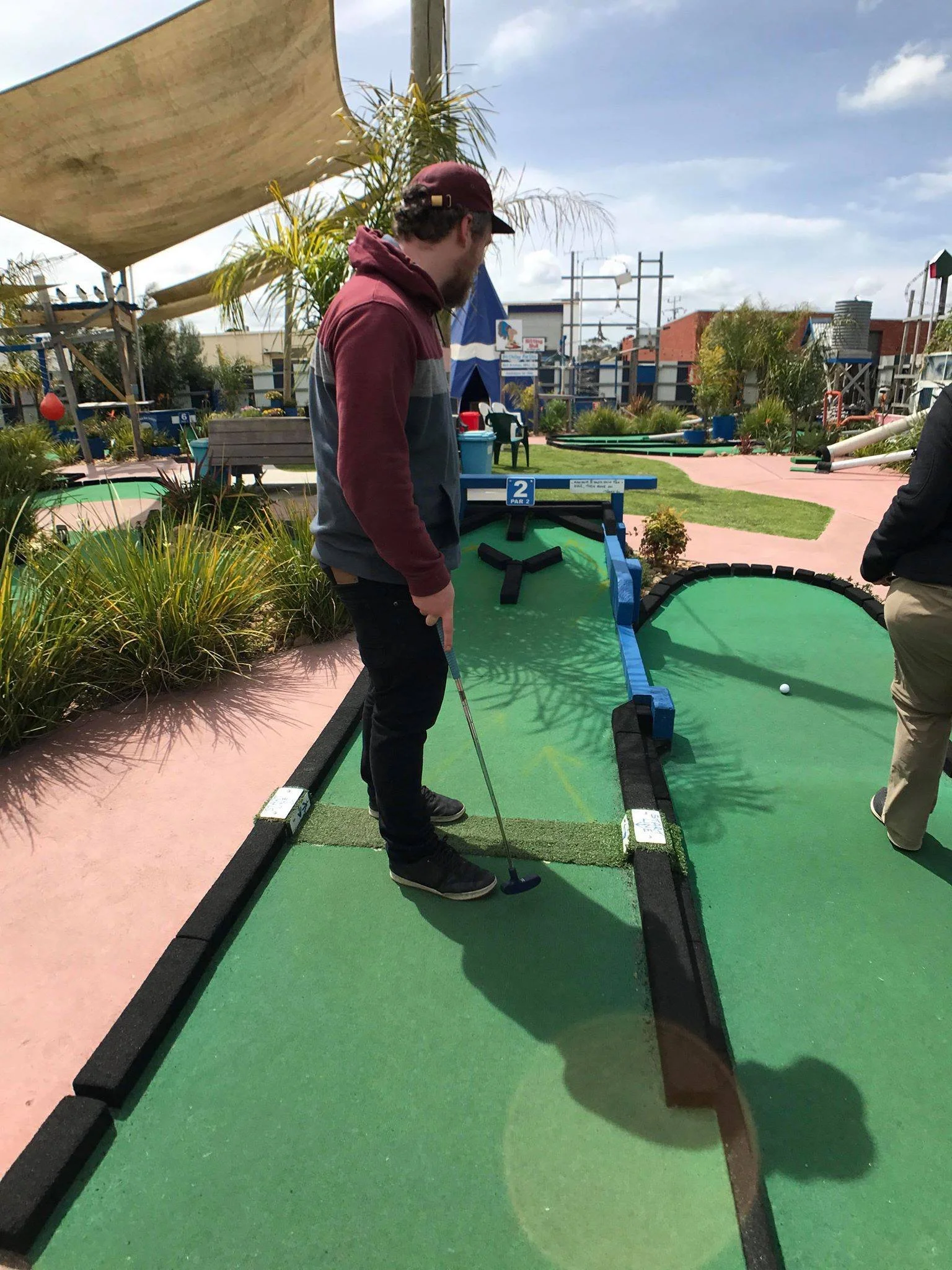 A man playing mini golf, standing over the hole on a green artificial turf course outdoors on a partly cloudy day.