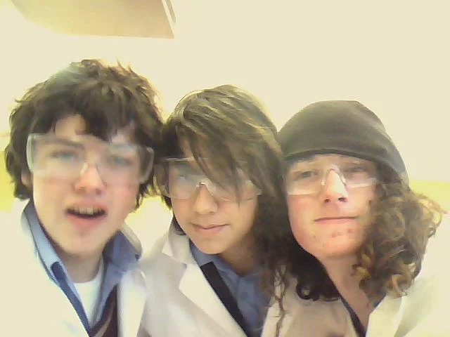 Three young men wearing safety glasses and lab coats taking a selfie together.