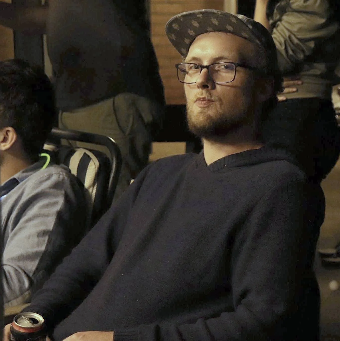 Young man wearing glasses, a black hoodie, and a patterned cap, sitting and looking at the camera in a social setting with other people around.