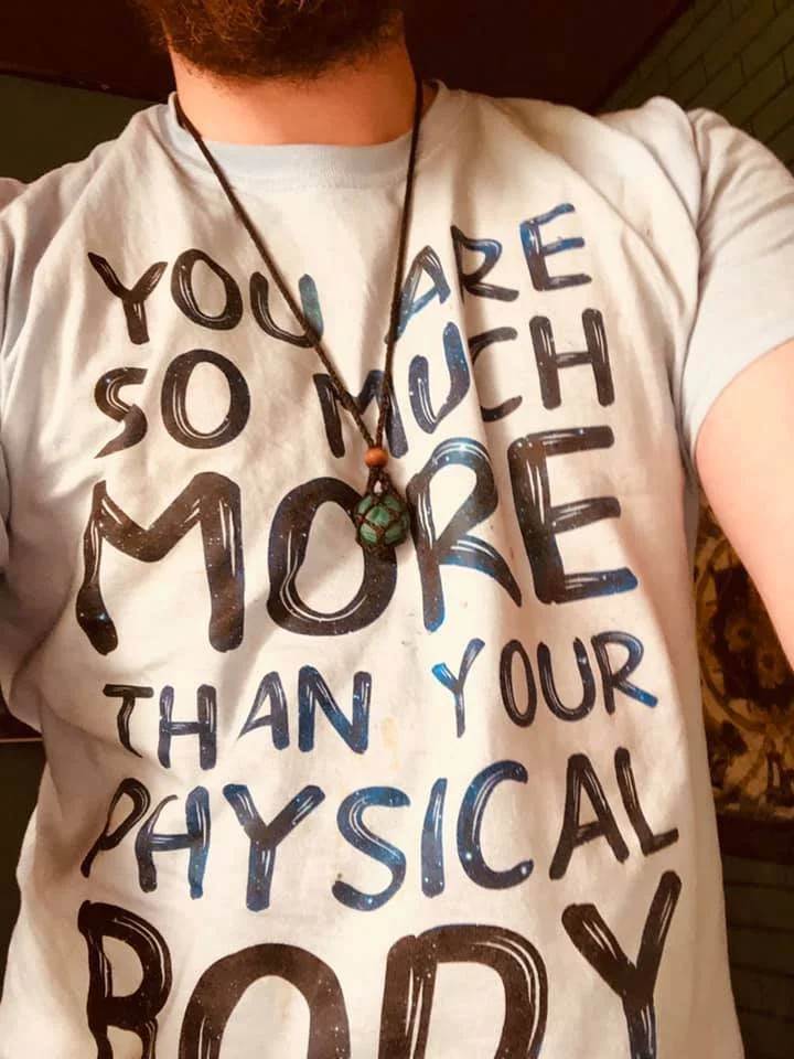 Person wearing a light-colored T-shirt with the words 'You are so much more than your physical body' written on it, and a necklace with a green snack and a bead.