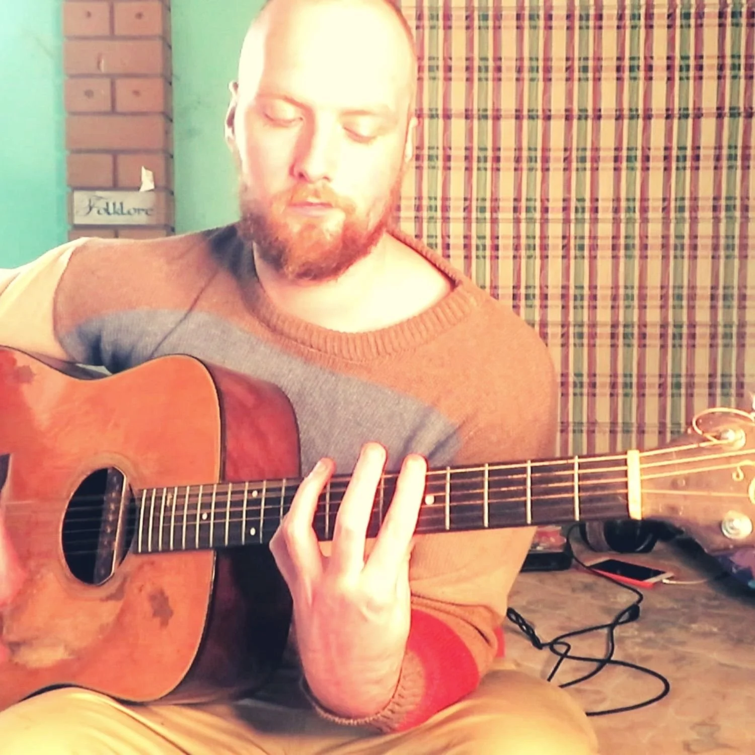 A man with closed eyes and a beard playing an acoustic guitar indoors, sitting on the floor with a patterned curtain in the background.
