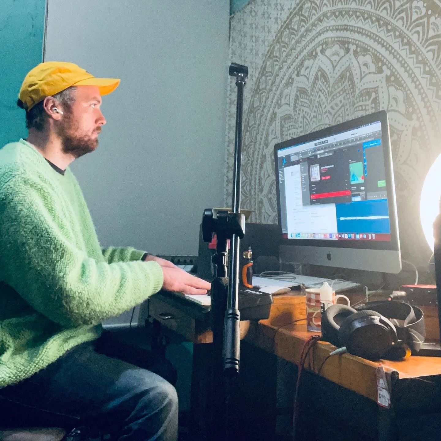 A man wearing a yellow cap and green sweater is sitting at a desk using a small keyboard, with a large computer monitor displaying music production software. On the desk are headphones, a mug, and other electronic equipment.