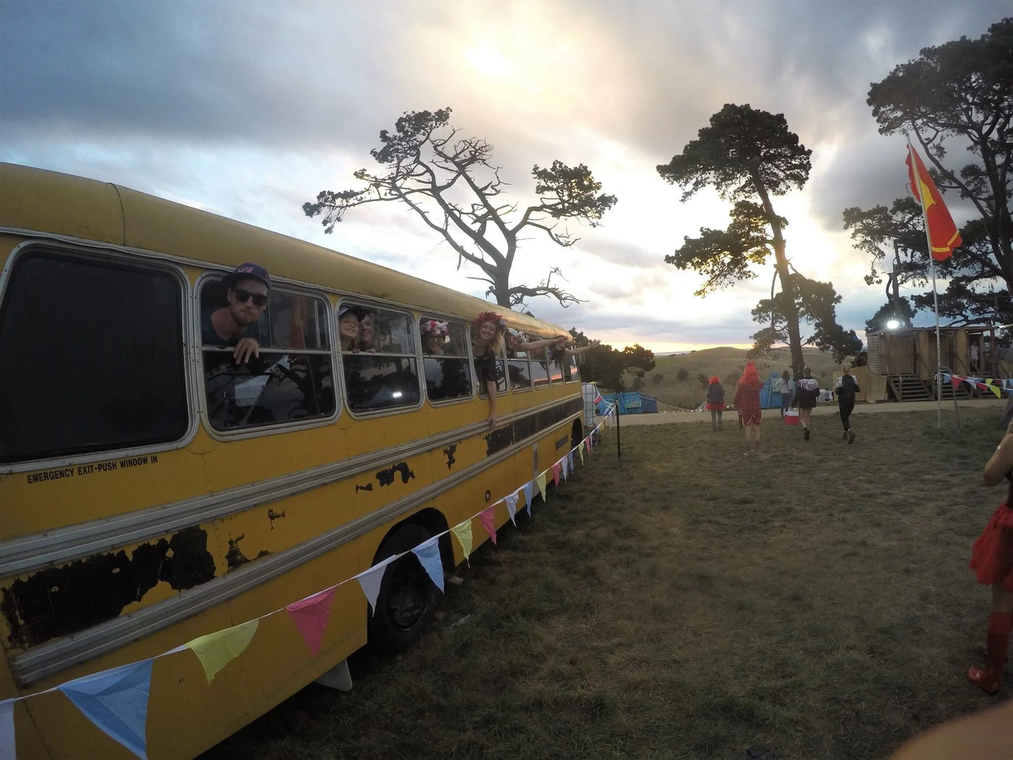 People inside an old yellow bus with peeling paint, looking out of the windows at sunset in a rural outdoor setting with trees and a field, some people walking around, flags, and tents in the background.
