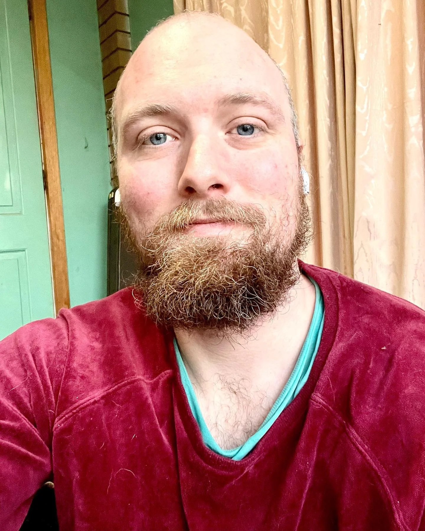 A man with a beard and blue eyes taking a selfie indoors, with green and beige curtains in the background.