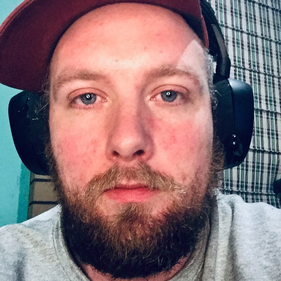 A close-up of a man with a beard wearing a brown cap and large black headphones, with a plaid patterned background.