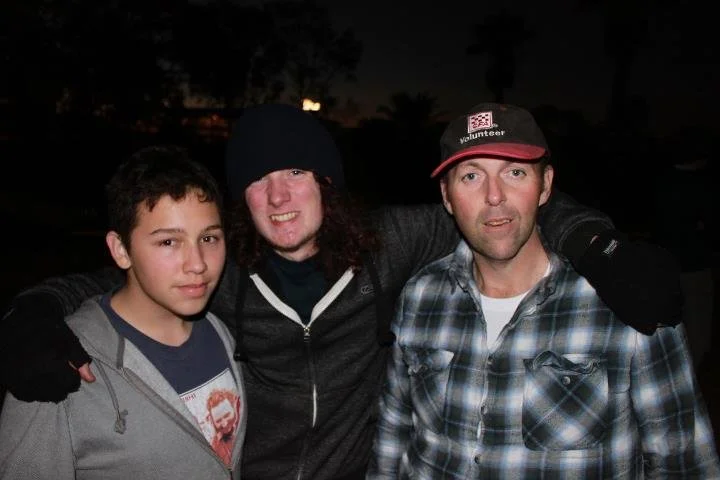 Three people standing outdoors at night, smiling with arms around each other.