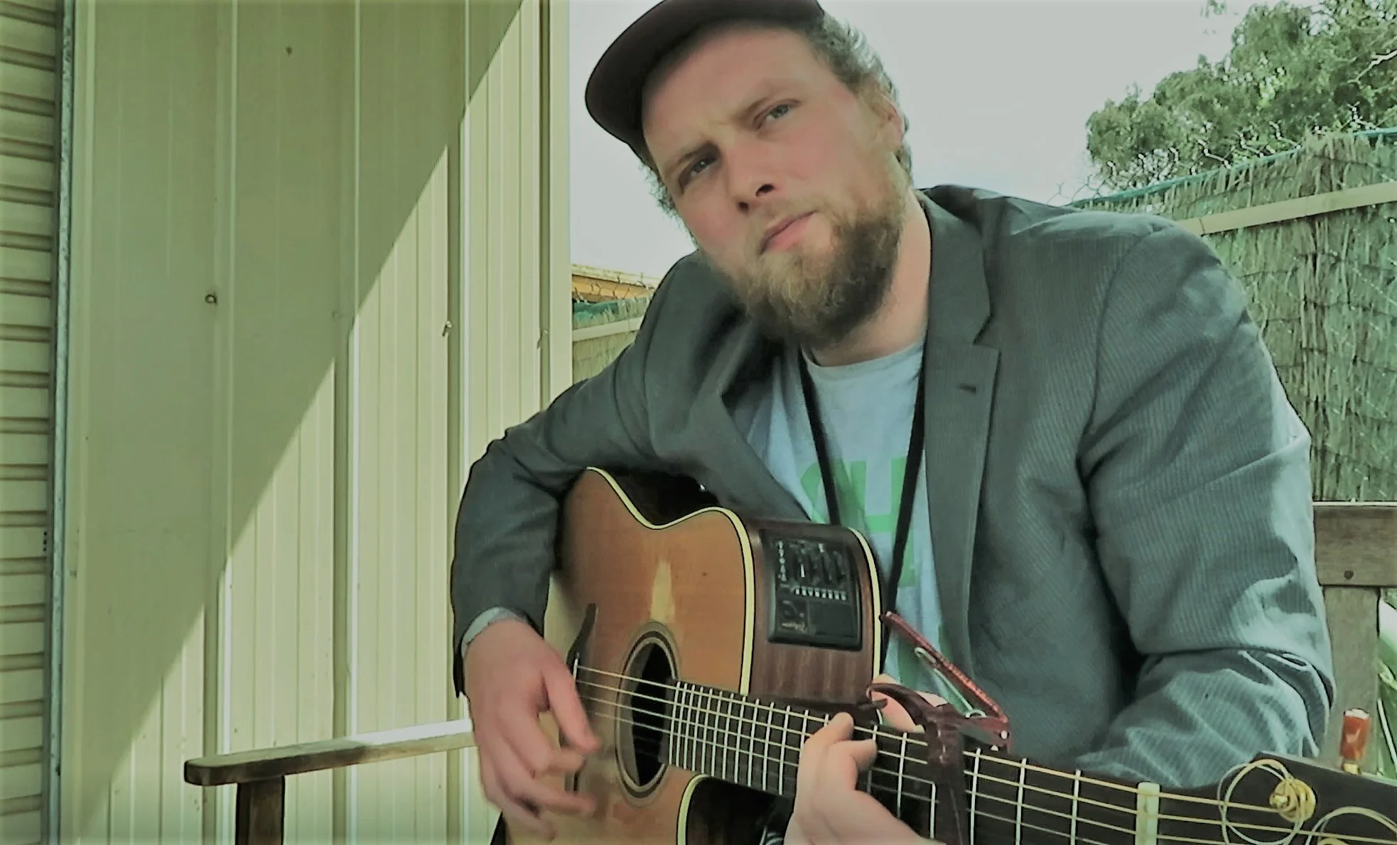 A man with a beard and mustache wearing a gray jacket and cap plays an acoustic guitar outdoors, near a light-colored metal wall and wooden fence.