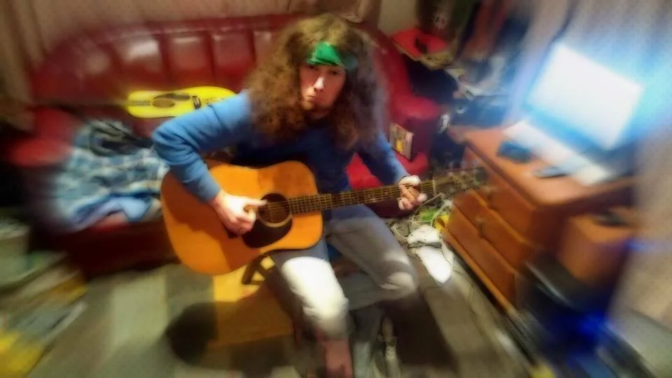 A person with long, curly hair wearing a green headband and a blue shirt sitting on a wooden chair, playing an acoustic guitar in a room with a red couch, a wooden entertainment center, and a computer.