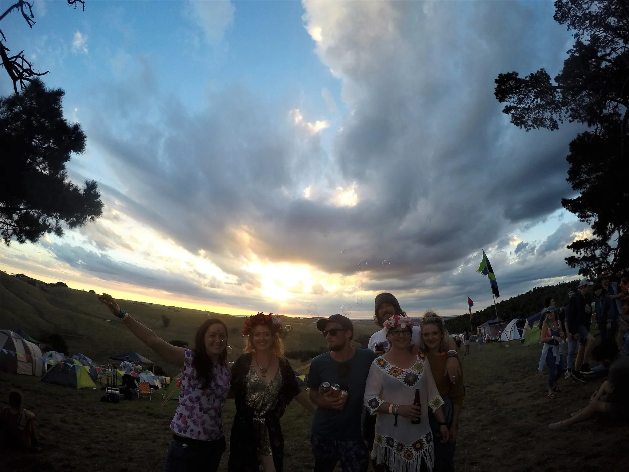 Group of six people standing outdoors during sunset, smiling and celebrating, with tents and flags in the background, under a partly cloudy sky.