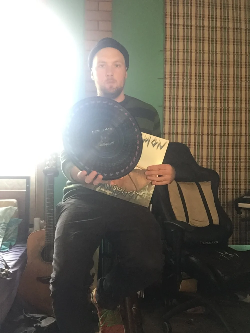 A man sitting on a chair holding a disc golf disc in a room with a guitar, a backpack, and a bed visible in the background.