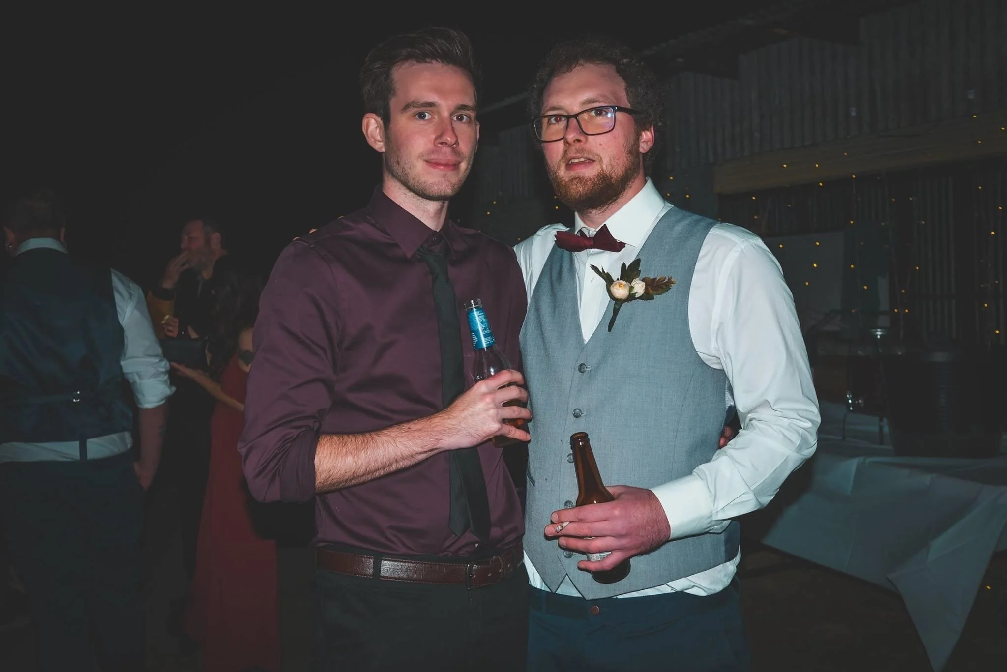 Two men at a social event holding drinks, wearing formal attire, with other guests in the background.