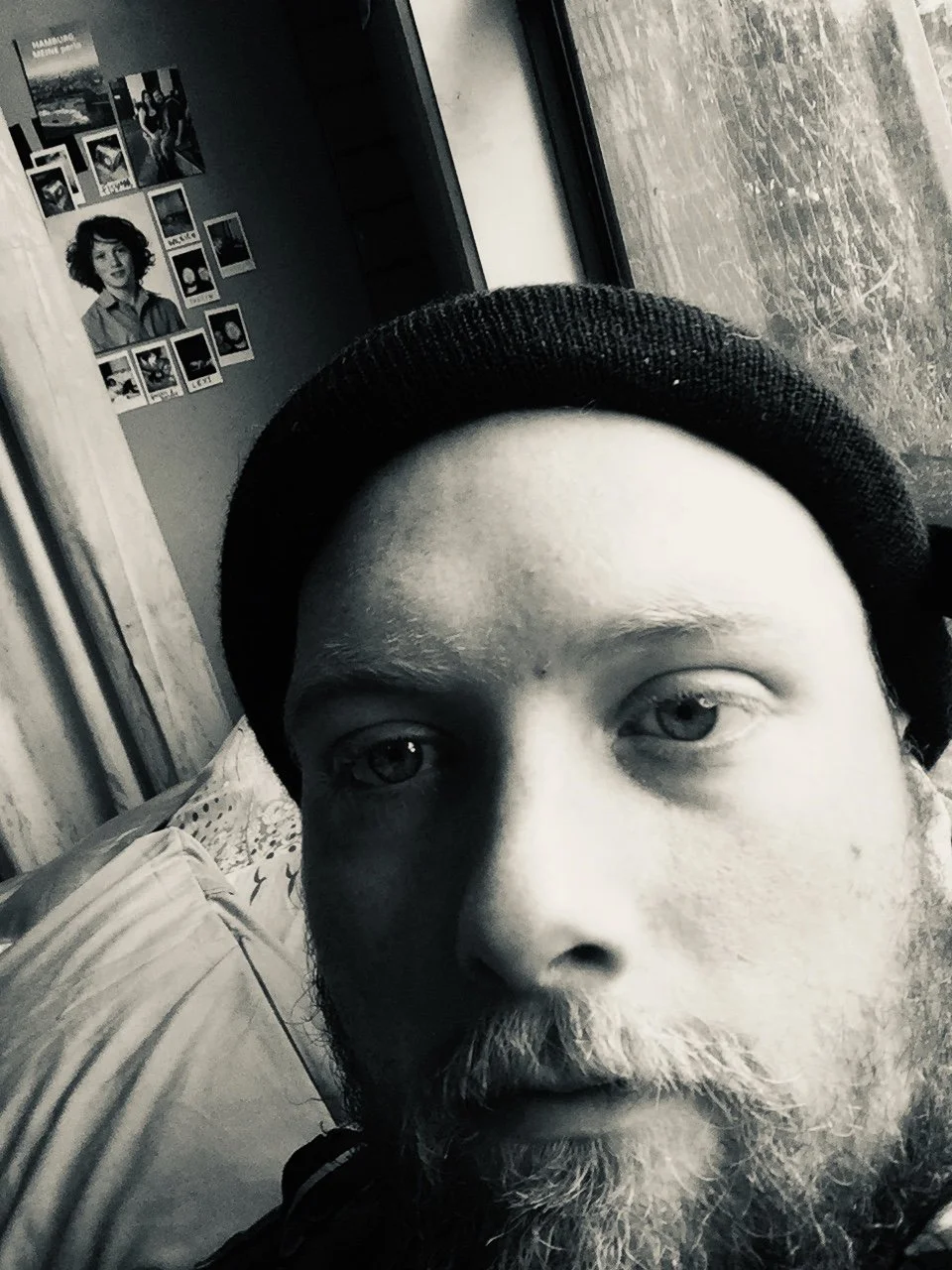 A black-and-white close-up selfie of a man with a beard wearing a beanie, taken indoors near a window with a tree outside. A wall with a collage of photos is visible in the background.