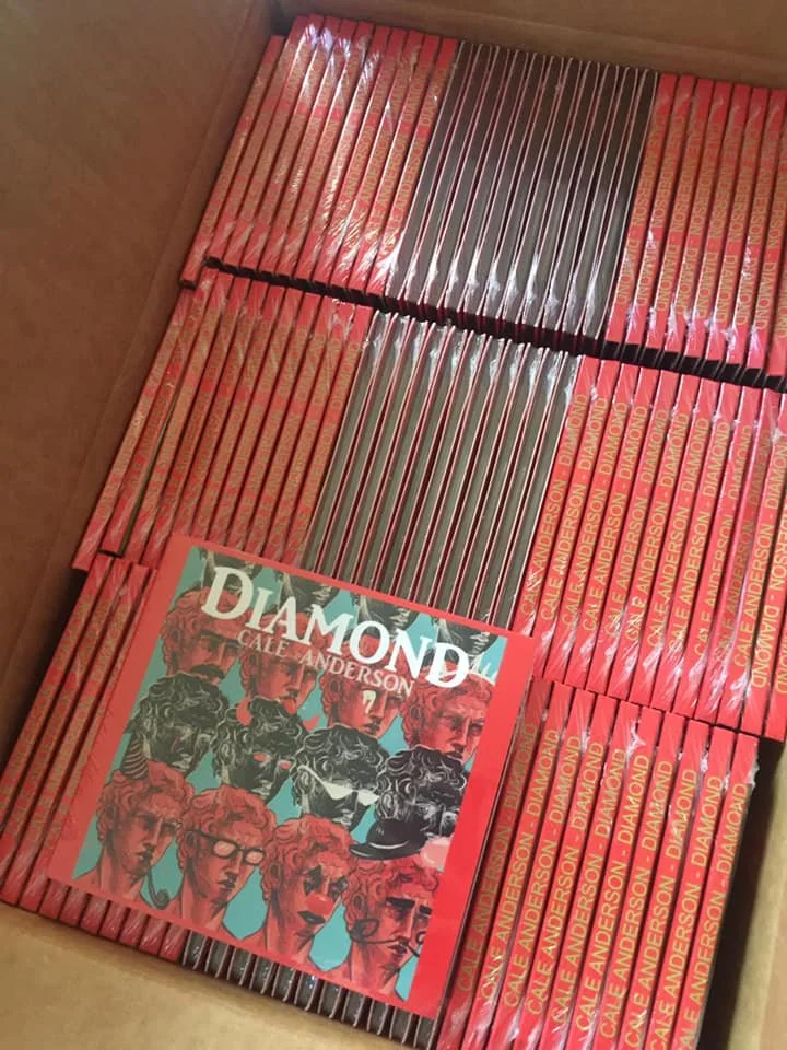 Box filled with multiple copies of the book titled 'Diamond' by Cale Anderson, featuring a colorful cover design with various illustrated faces.