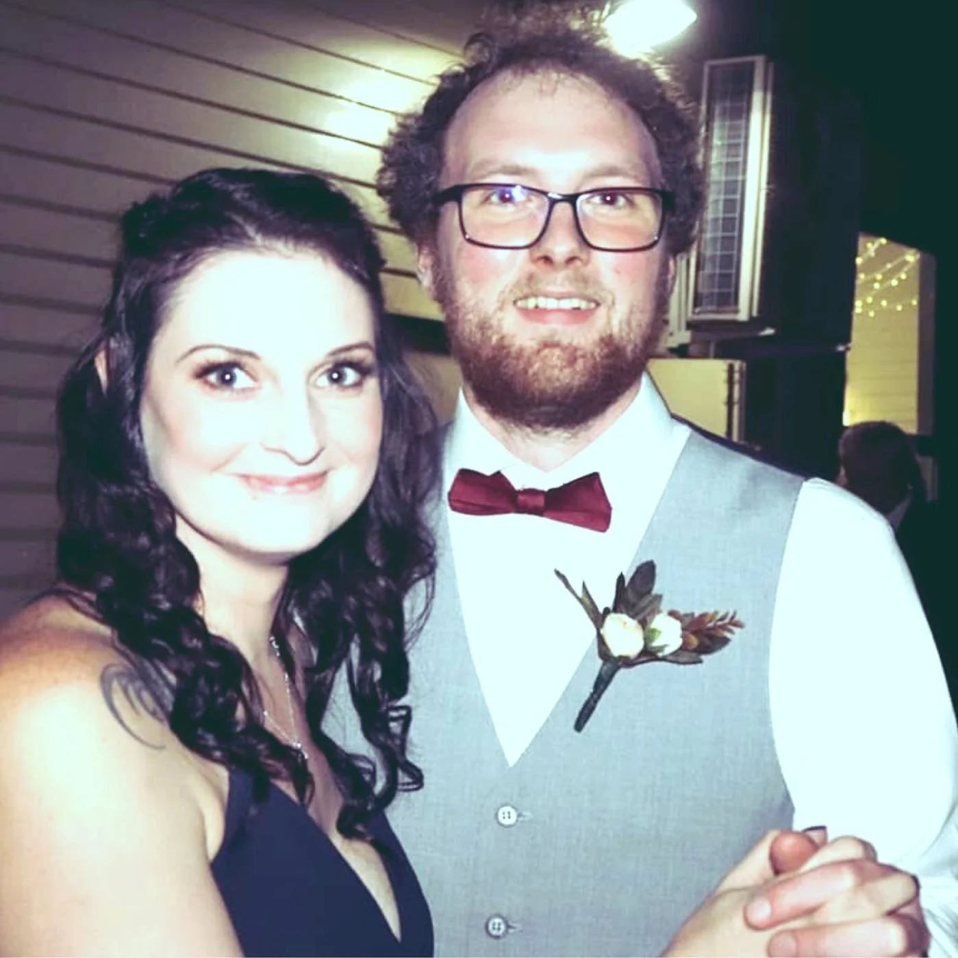 A woman with dark, curly hair and a man with glasses and a beard, dressed in formal attire, smiling together at a wedding or special event.