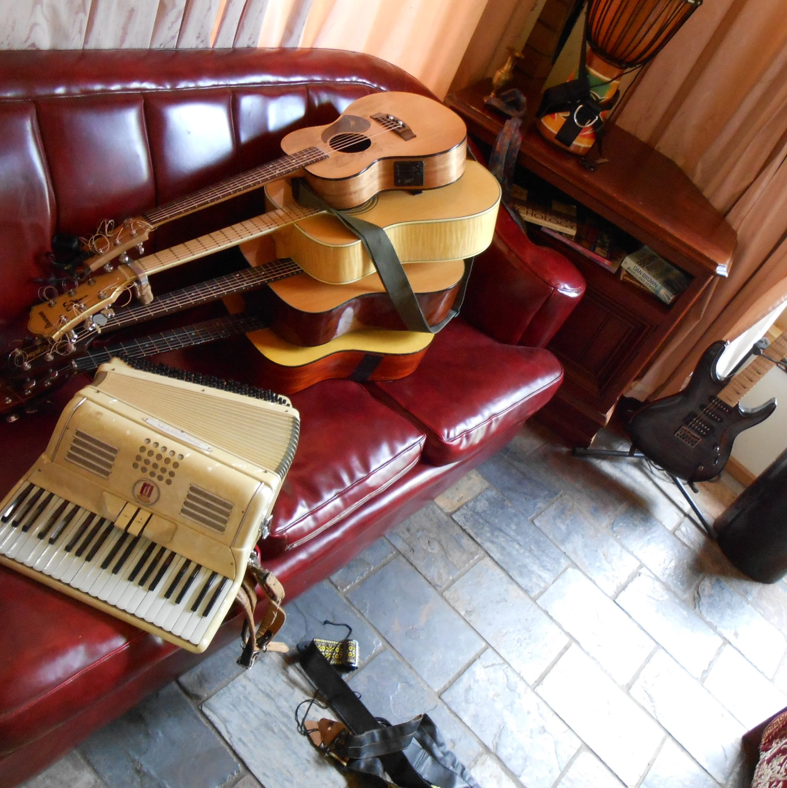 Assorted musical instruments including four guitars stacked on a red leather couch, a small keyboard, and an electric guitar on a stand next to a wooden cabinet in a room with tiled flooring.