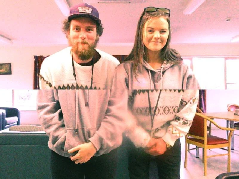 A man and a woman stand side by side in a brightly lit room, both wearing casual hooded sweatshirts. The man has a beard, curly hair, and a cap, while the woman has long straight hair and sunglasses on her head.
