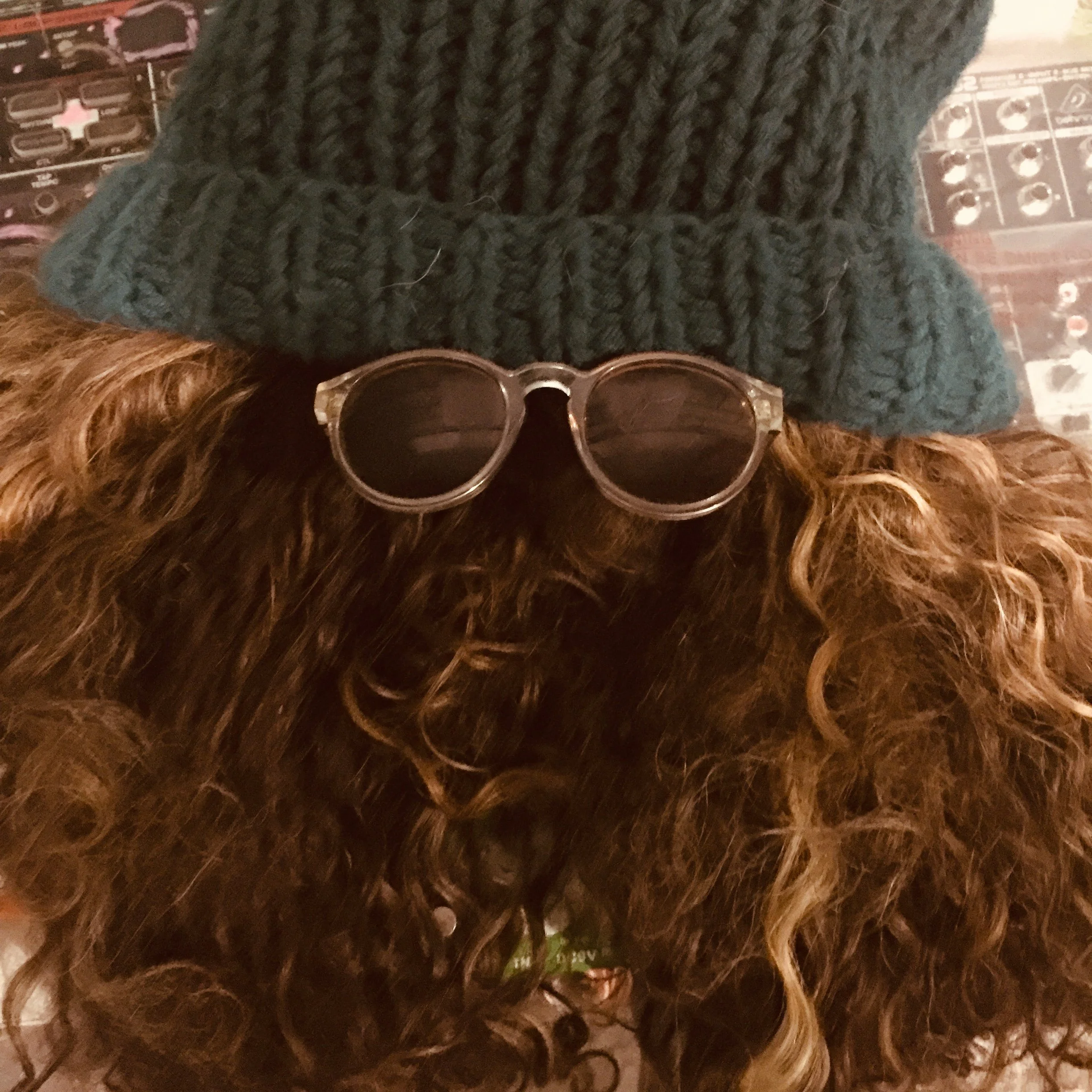 A person with curly brown hair wearing a green knit hat, sunglasses, and a green shirt.