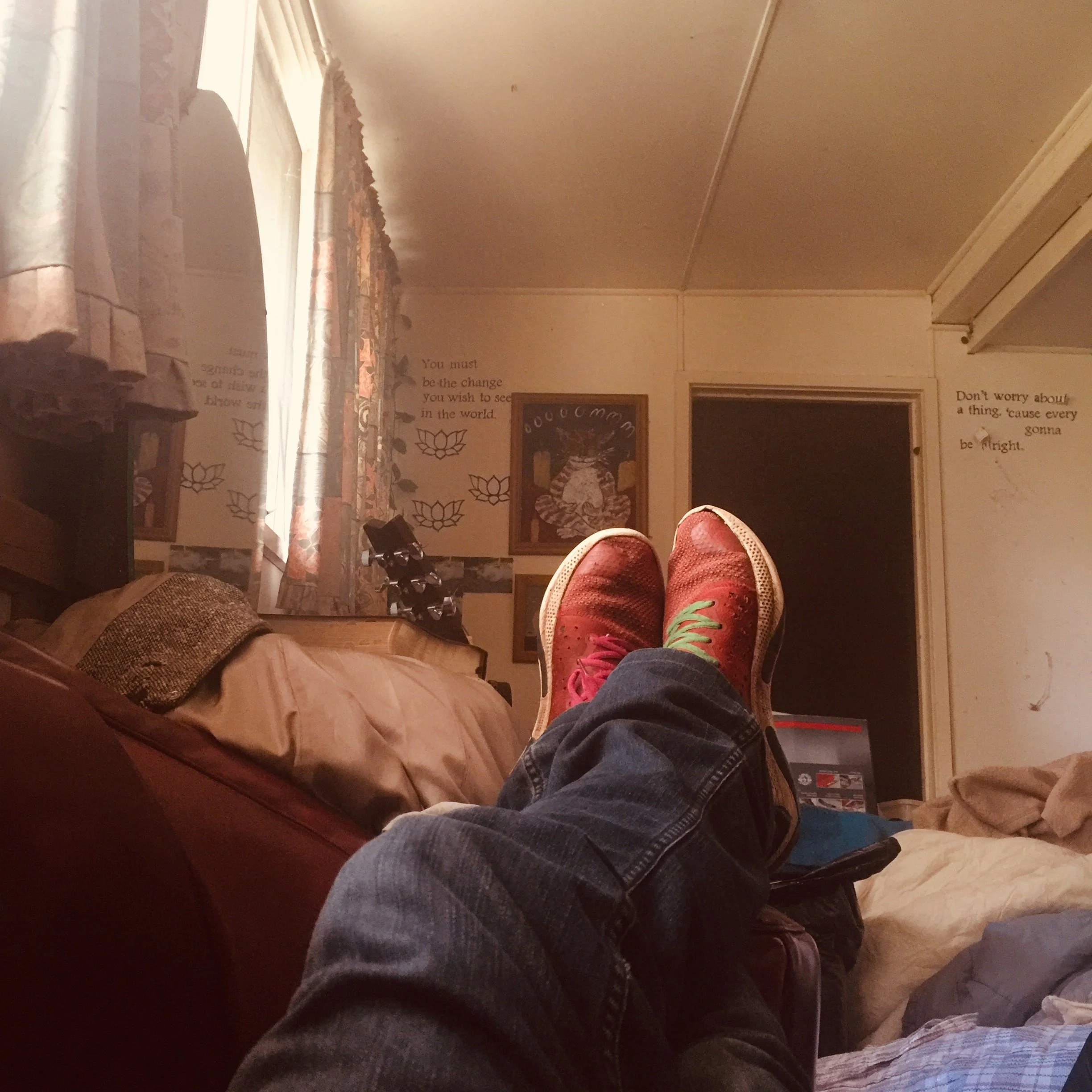 Person with crossed legs wearing red sneakers and jeans, relaxing on a couch in a living room with curtains, wall decorations, and a doorway in the background.