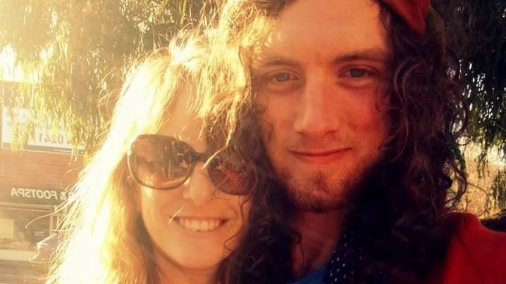 Two young adults, a woman with curly blonde hair and sunglasses and a man with long curly hair, are smiling outdoors during golden hour, with sunlight illuminating their faces.